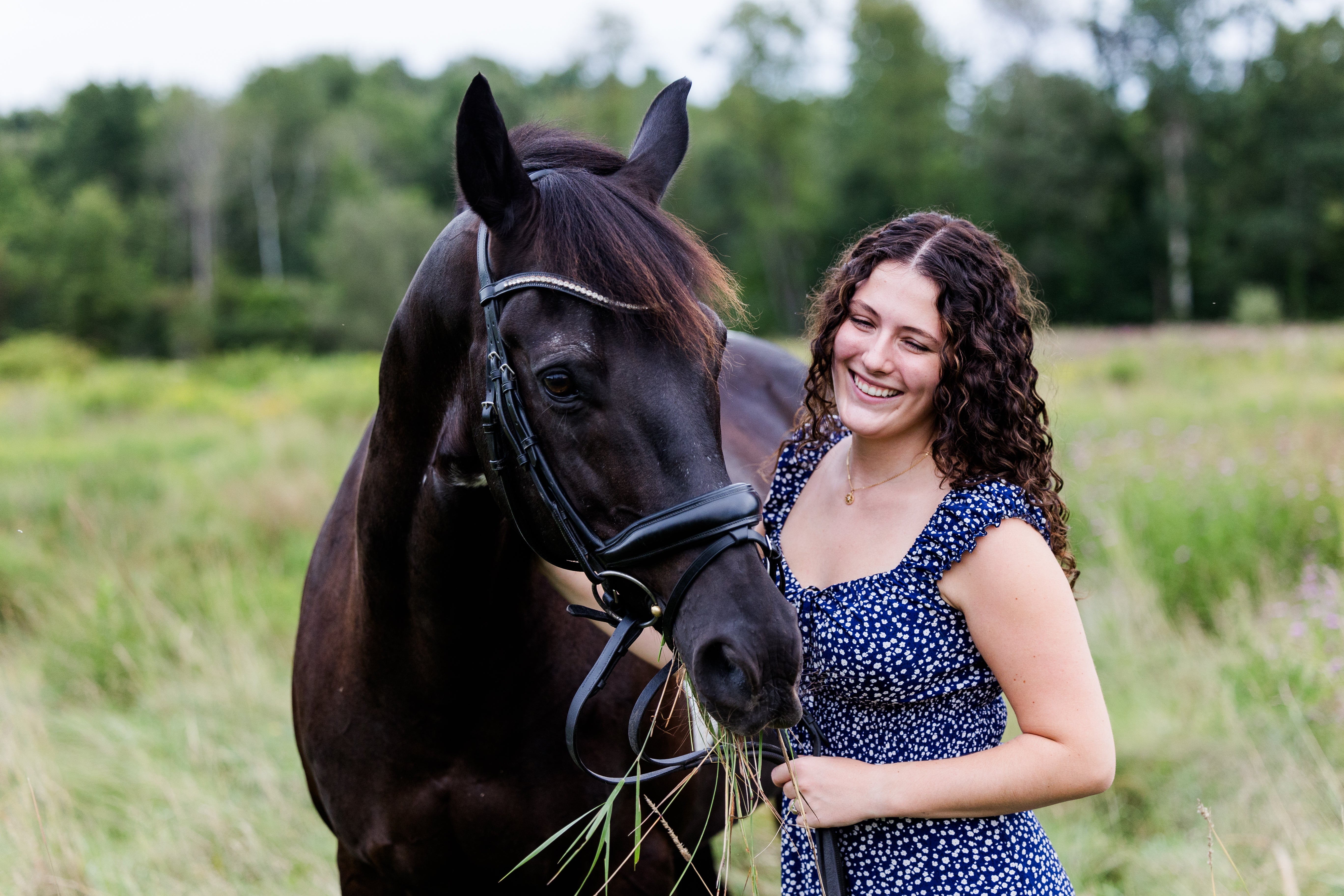 girl smiles in blue dress while laughing at horse eating grass